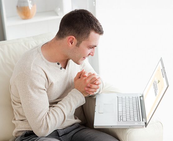 A man in a light-colored shirt bowing his head over a laptop keyboard with hands clasped in prayer, illustrating the prayer submission functionality and a user engaging with the spiritual service online.