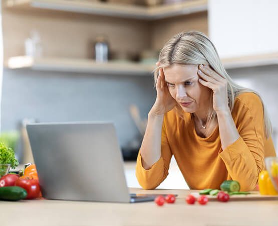 A woman sitting in a kitchen, holding her head in an expression of frustration or stress, symbolizing the complexity and challenges of managing dietary and health issues like diabetes.